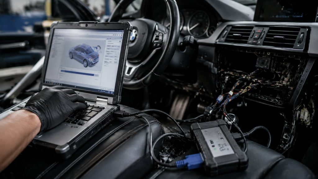 Automotive technician performing ECU programming and J2534 pass-through reflash on vehicle computer module using diagnostic laptop in workshop